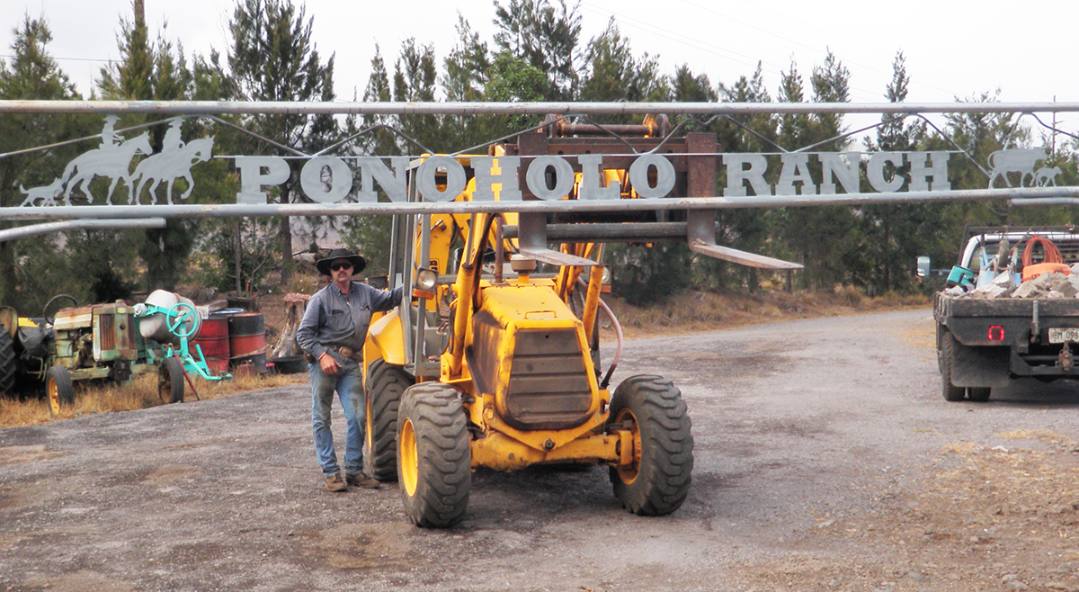 Jeff Hanneken stands with yellow JCB tractor holding Ponoholo Ranch metal sign